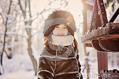Happy adorable child girl in fur hat and coat near bird feeder on the walk in winter forest Stock Photo