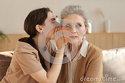 Happy adult daughter whispering secret to her middle mother at home, gossiping, sharing secret Stock Photo