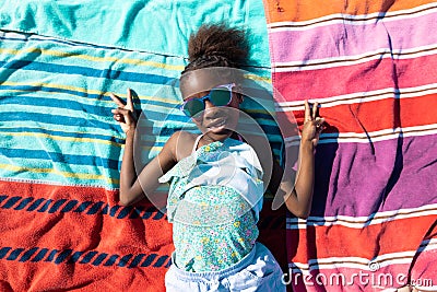 Happy african american girl in sunglasses showing peace sign while lying on towels at beach Stock Photo