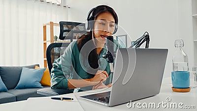 Happy asia girl record a podcast on her laptop computer with headphones and microphone talk with audience at her room. Female Stock Photo