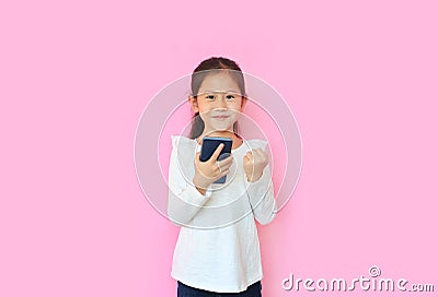 Happy asian little child girl watching her smartphone and expression hand fight sign over pink background. Kid smiling and enjoy Stock Photo