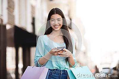 Happy Asian Shopper Girl Posing Outdoors With Smartphone And Shopping Bags Stock Photo