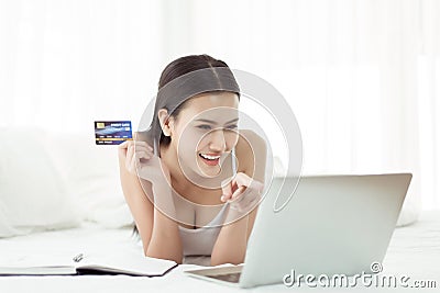 Happy Asian woman holding mockup credit card for shopping online and finger pointing to laptop in bedroom Stock Photo