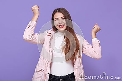Happy attractive girl enjoys every moment of life, raises arms and clenches fists, looks at camera, has good mood, wears pale pink Stock Photo