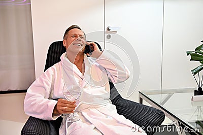 Happy attractive mature European man with a drinking glass with water in hand, sitting on an armchair in wellness spa lounge zone Stock Photo