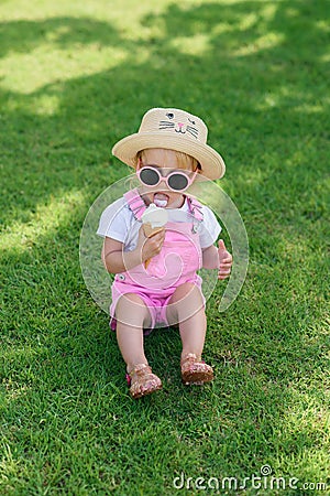 Happy baby girl dressed pink summer clothes, yellow hat and pink sunglasses sits on a green lawn and eats white ice Stock Photo
