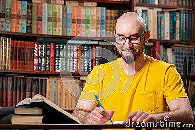 A happy bald, bearded man with glasses is writing something in a notebook. There are bookshelves in the background. Concept of Stock Photo