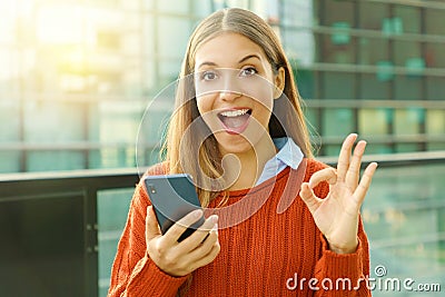 Happy beautiful autumn woman holding smart phone doing Ok sign at the camera. Excited satisfied young woman using smart phone Stock Photo