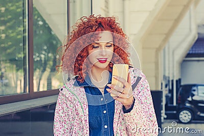 Happy beautiful woman reading good news message or seeing photos on her mobile phone. Smiling red head curly hair girl, isolated Stock Photo