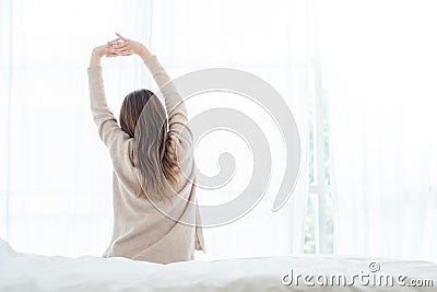 Happy beautiful young Asian woman waking up in morning, sitting on bed, stretching in cozy bedroom, looking through window. Stock Photo