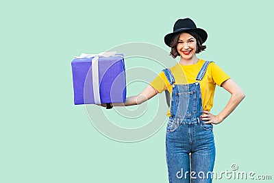 Happy beautiful young girl in hipster wear in denim overalls and black hat standing and holding big heavy gift box with toothy Stock Photo