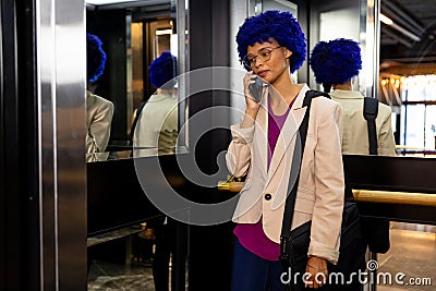 Happy biracial casual businesswoman with blue afro talking on smartphone in office elevator Stock Photo