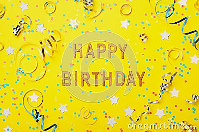 Happy birthday greeting text decorated with confetti and serpentine on yellow background top view. Flat lay style. Stock Photo