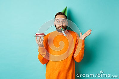 Happy birthday guy celebrating, wearing party hat, blowing wistle and holding bday cake, standing against white Stock Photo