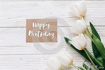 happy birthday text sign on stylish craft greeting card and tulips on white wooden rustic background. flat lay with flowers and g Stock Photo