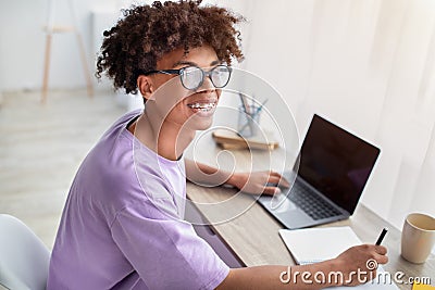 Happy black adolescent sitting at desk with laptop, learning remotely, taking notes in copybook at home Stock Photo