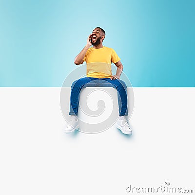 Happy black man sitting on white blank placard, shouting, announcing huge sale on blue background, mockup Stock Photo