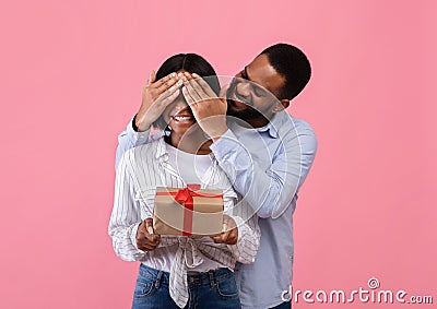 Happy black woman holding Valentine`s gift box, loving boyfriend covering her eyes on pink studio background Stock Photo
