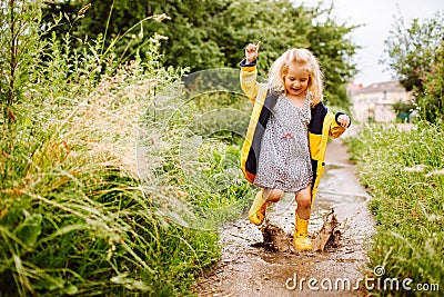 Happy blonde child girl in a yellow rubber boots and yellow raincoat jumping in a puddle. Summer, childhood, holiday Stock Photo