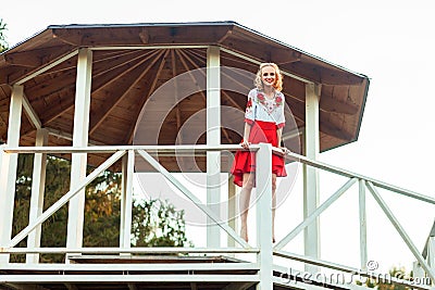 Happy blonde young woman in stylish red white dress posing in wooden garden arbor. standing and looking at camera with toothy Stock Photo
