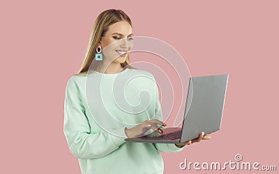 Happy blondy woman in mint sweatshirt doing remote job working on laptop on pink background. Stock Photo