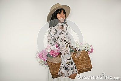 Happy boho girl smiling and holding pink and white peonies in rustic basket. Stylish hipster woman in hat and bohemian floral Stock Photo