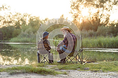 Happy boy sitting on chairs together with his experienced old grey-bearded grandpa and fishing with rods on the lake. Stock Photo
