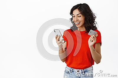 Happy brunette girl holds credit card and looks pleased at smartphone screen, checks bank account finance app, purchase Stock Photo