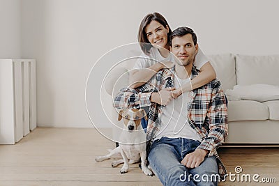 Happy brunette woman embraces husband with love, being in good mood, smiles positively. Husband, wife and dog pose together in Stock Photo
