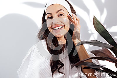 Happy brunette woman in a white bathrobe takes care of her skin and moisturizes her face with a cream Stock Photo