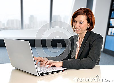 Happy business woman with red hair smiling at work typing on computer laptop at modern office desk Stock Photo