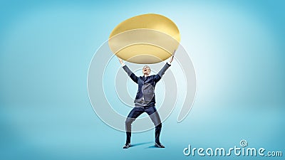A happy businessman on blue background holds a huge golden egg over his head. Stock Photo