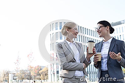 Happy businesswomen conversing while holding disposable cups outside office building Stock Photo