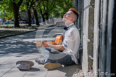 Happy busker street musician sitting on a city sidewalk and playing music with guitar. Freedom, music and art concept Stock Photo