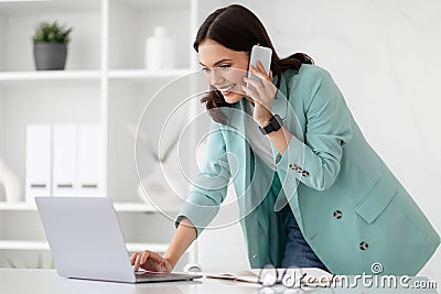 Happy busy millennial caucasian lady in suit typing on computer, calls by phone at workplace Stock Photo