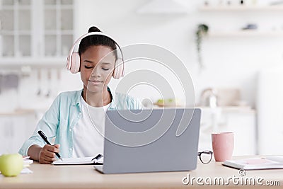 Happy busy teen black girl in earphones studying, doing homework at table with laptop in kitchen interior Stock Photo