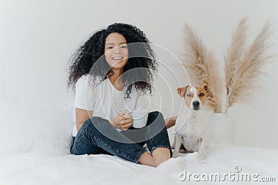 Happy carefree woman with Afro hairstyle, wears t shirt nad jeans, feels glad, holds mug of coffee, laughs sincerely, jack russell Stock Photo