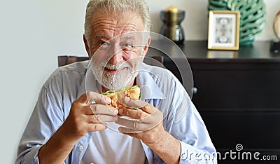 Happy caucasian elderly eating hamburger in living room with smiling face Stock Photo