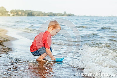Happy Caucasian red-haired toddler child boy putting blue paper boat in water on lake sea ocean Stock Photo