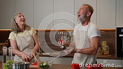 Happy Caucasian senior old couple having fun cooking at home funny man fooling around juggling tomatoes smiling woman Stock Photo