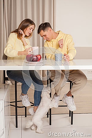 Happy cherrful couple in the kitchen sitting by the table and drinking from the cups, dog under the table Stock Photo