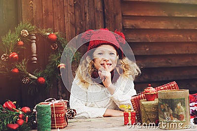Happy child girl in red hat and scarf wrapping Christmas gifts at cozy country house, decorated for New Year and Christmas Stock Photo