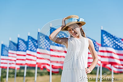 Child standing in white dress and touching straw hat near american flags Stock Photo