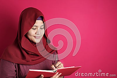 Happy confident successful Asian muslim woman wearing hijab writing on book, smiling and thinking Stock Photo