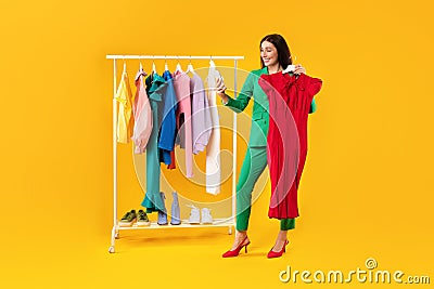 Happy woman shopaholic using smartphone and holding hanger with new dress and taking selfie, posing near clothing rail Stock Photo