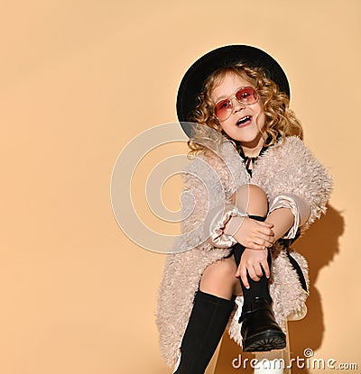 Little girl in sunglasses, pink fur coat, black hat and boots. Looking surprised, sitting cross-legged against beige background Stock Photo