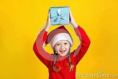 Happy cute baby holds a box over his head. Little girl in santa claus hat with christmas present on yellow background Stock Photo