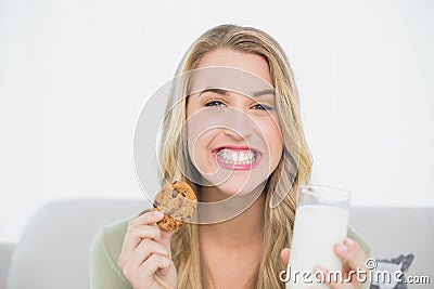 Happy cute blonde eating cookie with milk sitting on cosy sofa Stock Photo