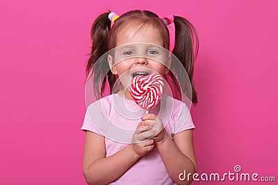 Happy cute girl wears rose t hirt, stands isolated over pink background, holds bright lollipop in hands. Cheery child with opened Stock Photo