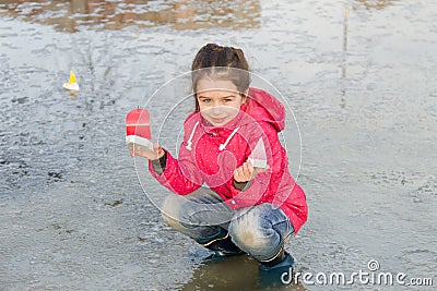 Happy cute little girl in rain boots playing with handmade colorful ships in the spring creek standing in water Stock Photo